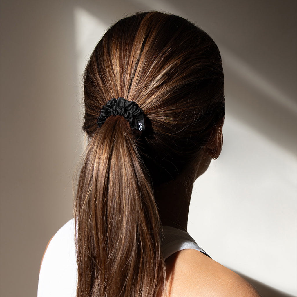 A person with long, straight brown hair is seen from behind, wearing a white sleeveless top and a black scrunchie holding their hair in a low ponytail, with soft light and shadows on the wall.