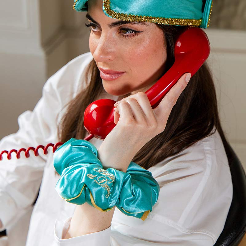 Wearing a turquoise "Breakfast at Drowsy" Padded Scrunchie by drowsysleepco, a woman in a white shirt and matching sleep mask holds a red vintage phone, her voluminous hair styled elegantly indoors as she gazes thoughtfully to the side.