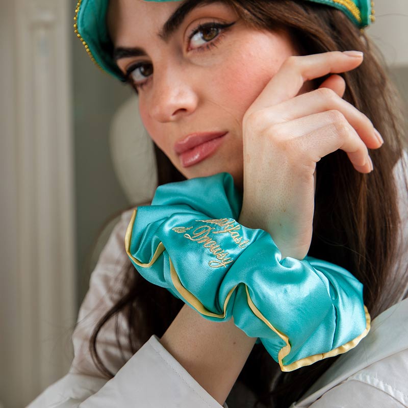 A woman rests her chin on her hand, wearing the drowsysleepco Hollywood Experience Bundle—an eyelash-protecting teal silk scrunchie with gold trim and matching sleep mask, both featuring embroidered text.