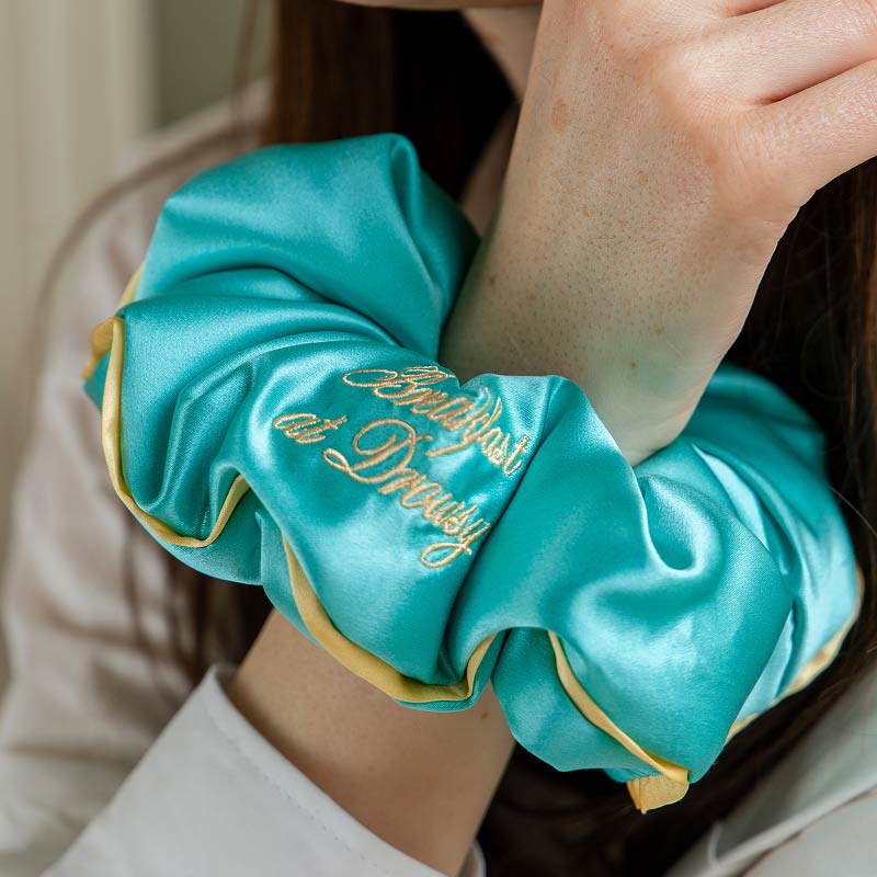 A person wears a large, turquoise Breakfast at Drowsy - Turquoise Padded Scrunchie by drowsysleepco on their wrist; gold trim and embroidery visible. Their face is partially out of frame.