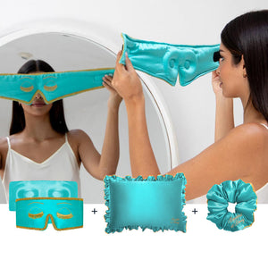 A woman in a white tank top holds drowsysleepco’s Hollywood Experience Bundle – Eyelash protecting, featuring a turquoise silk sleep mask with gold trim, matching eye mask, ruffled silk pillowcase, and coordinating scrunchie.