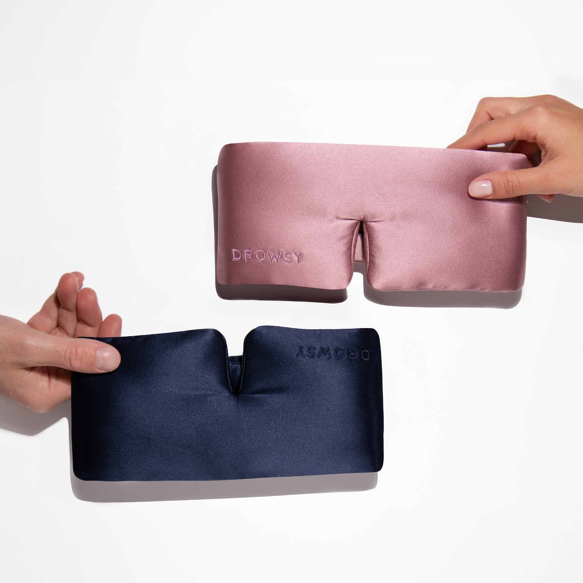Two hands each hold a silky, padded Drowsy Lovers Pack mask—one pink, one navy—crafted from mulberry silk for ultimate beauty sleep, both embroidered with DROWSY, against a plain white background.