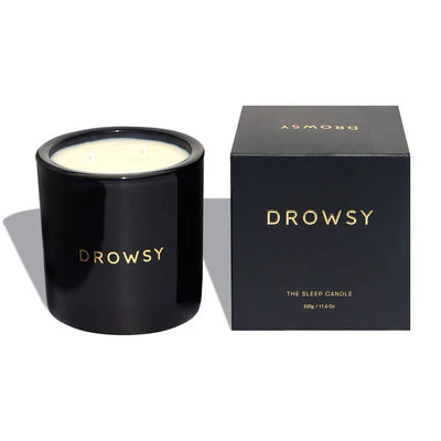 A black candle jar labeled Drowsy sits next to its matching black box, which also says Drowsy and The Sleep Candle in gold lettering. The candle appears unused, with two visible wicks.