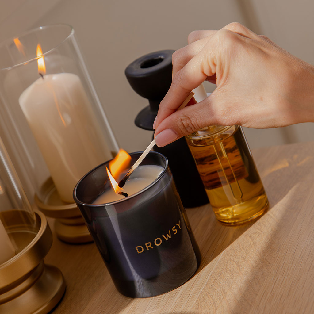 A hand lights a candle labeled DROWSY with a match. The candle sits on a wooden surface next to a glass bottle of oil, a black candle holder, and a large white candle in a glass container.