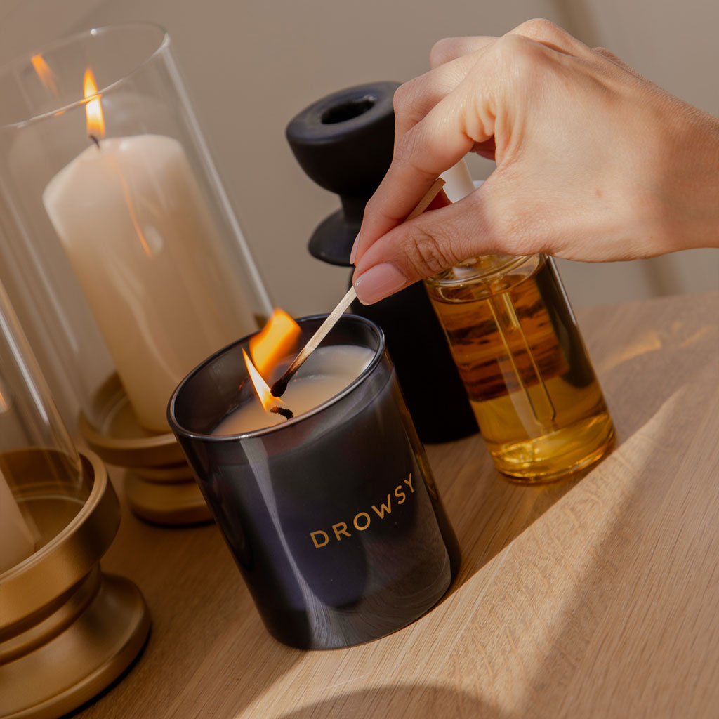 A hand lights the drowsysleepco Sleep Ritual Candle 220g, known for its calming scent, with a match. The candle rests on a wooden surface beside a glass oil bottle and decorative holders with lit and unlit candles.
