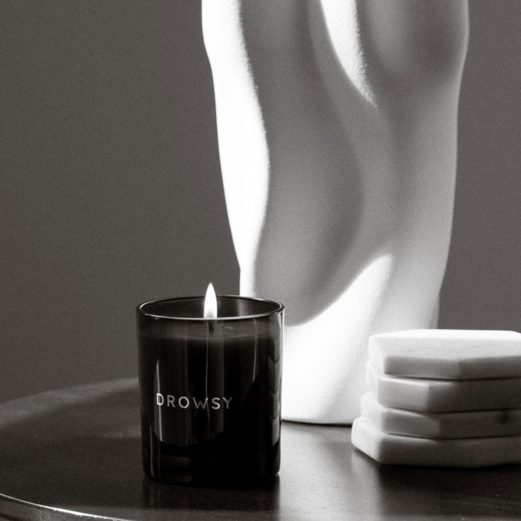 The Sleep Ritual Candle 220g from drowsysleepco burns on a round surface with a white abstract sculpture and stacked coasters, softly lit in black and white—ideal for adding relaxation to your pre-sleep routine.
