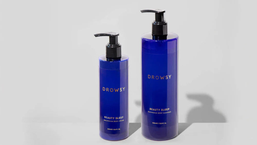 Two blue pump bottles labeled Drowsy Beauty Sleep Rejuvenating Body Cleanser are standing side by side on a light gray surface, with one bottle larger than the other.
