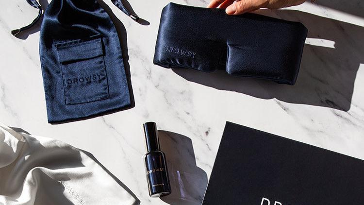 A hand holds a dark blue silk sleep mask labeled Drowsy on a marble surface, alongside a matching pouch, a spray bottle, a white cloth, and a black box, all branded with Drowsy.
