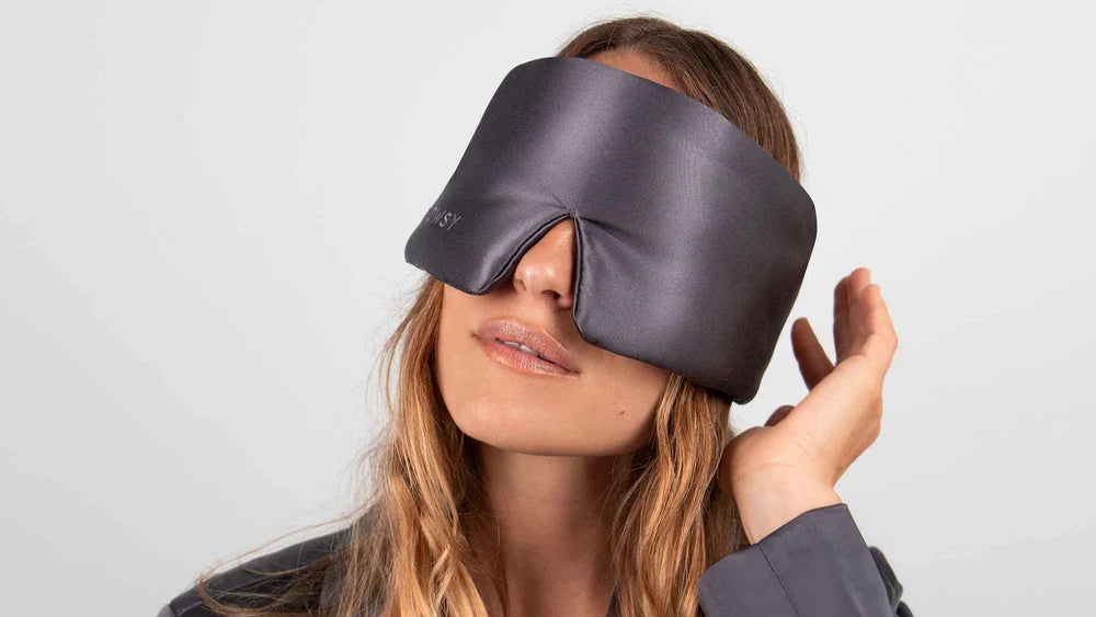 A woman with long hair wears a large, black, padded sleep mask that covers her eyes and part of her nose, while touching the edge of the mask with one hand.