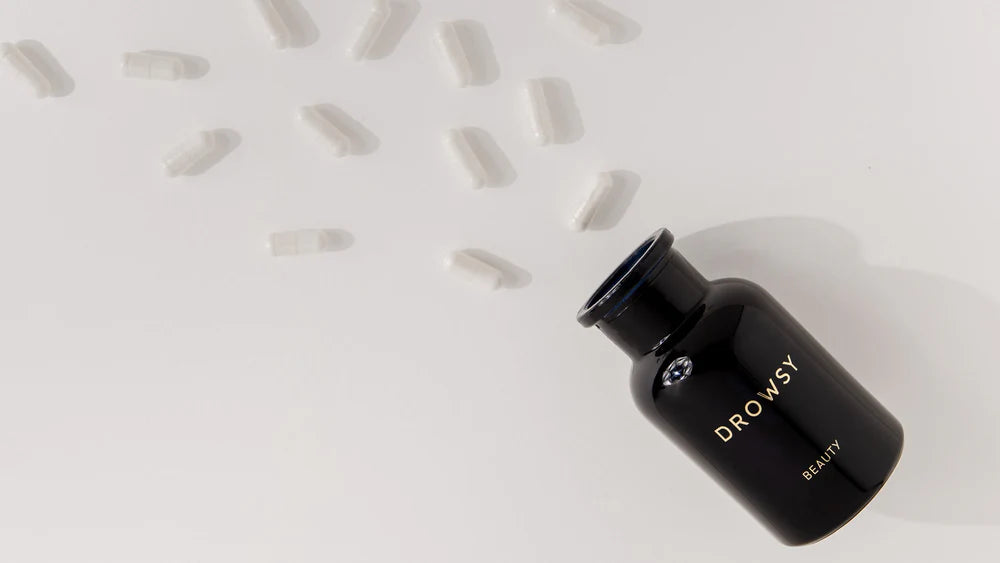 A black bottle labeled DROWSY BEAUTY lies on its side with white capsules scattered around it on a light surface.