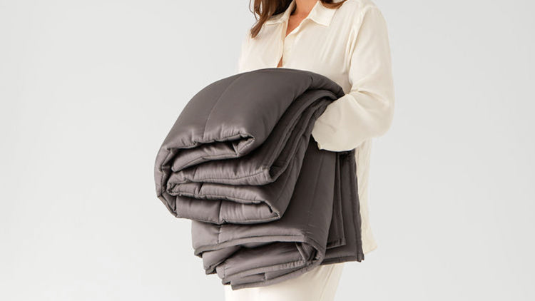 A person wearing light-colored pajamas holds a large, folded, dark gray weighted blanket against a plain white background.