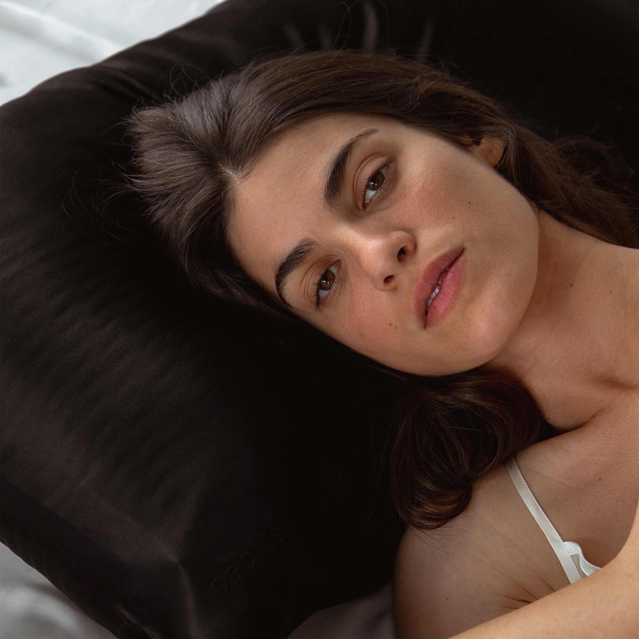 A woman with long brown hair rests on a Drowsy is the new black bundle silk pillowcase by drowsysleepco, gazing at the camera. Soft light enhances her calm look, capturing true beauty sleep.