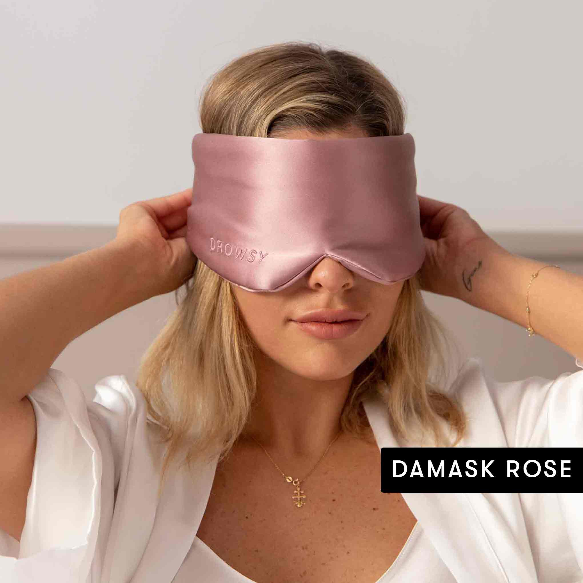 A woman in a white robe adjusts a silky pink Drowsy mask from the Besties Pack over her eyes, preparing for beauty sleep. DAMASK ROSE is displayed in the bottom right corner.