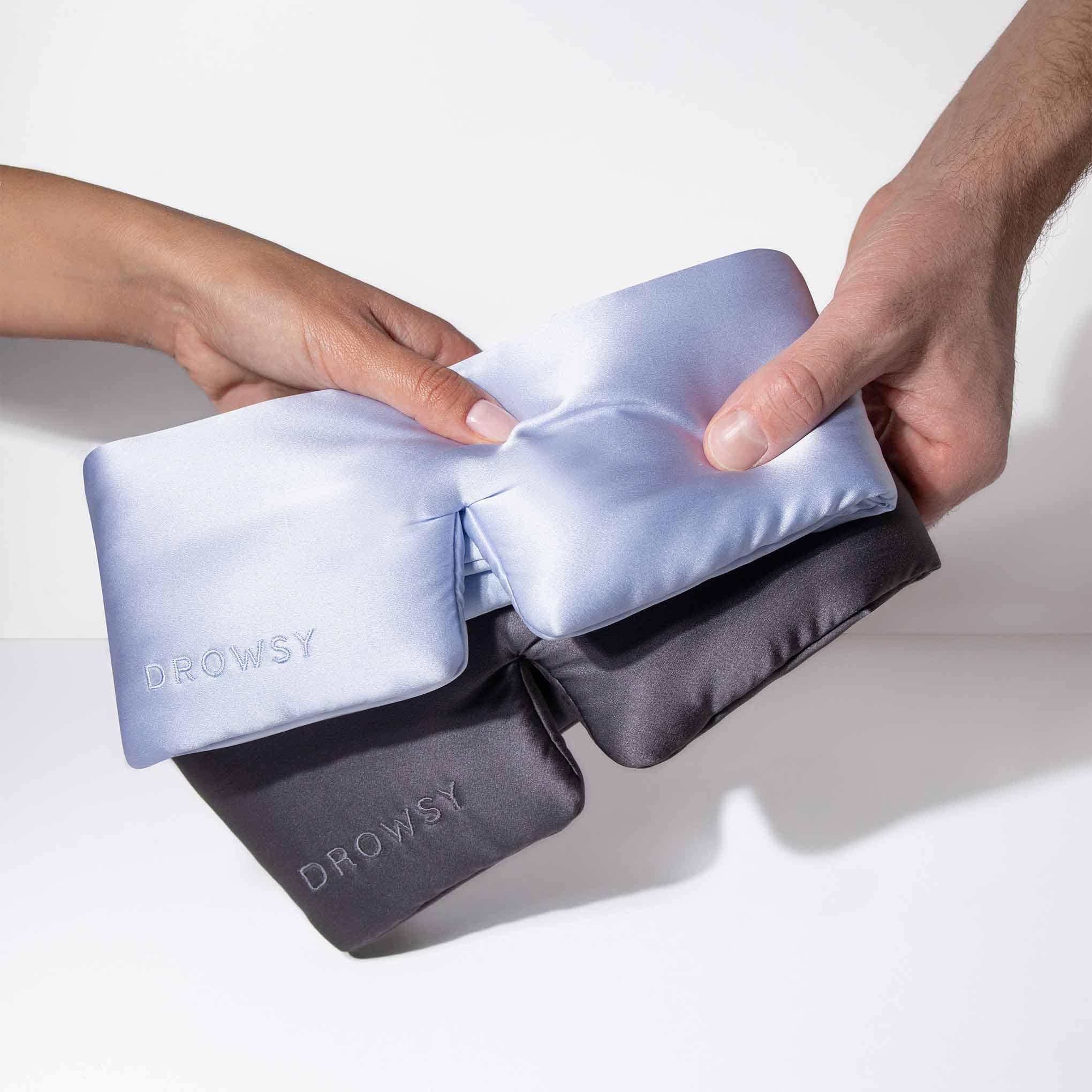 Two hands exchange Drowsy’s Lovers Pack—luxury silk masks in light blue and black, each embroidered “DROWSY”—promising beauty sleep with the finest mulberry silk against a plain white background.