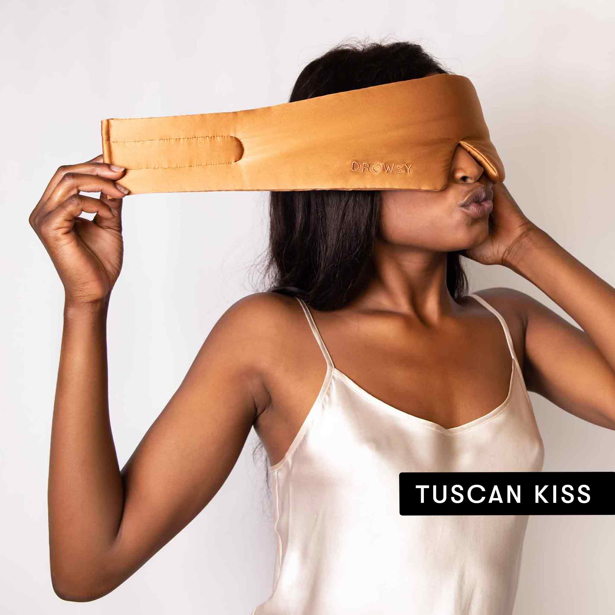 A woman in a cream satin camisole puckers her lips while wearing the Drowsy Besties Pack: a tan luxury silk mask, ideal for beauty sleep, elegantly presented in a black TUSCAN KISS box.