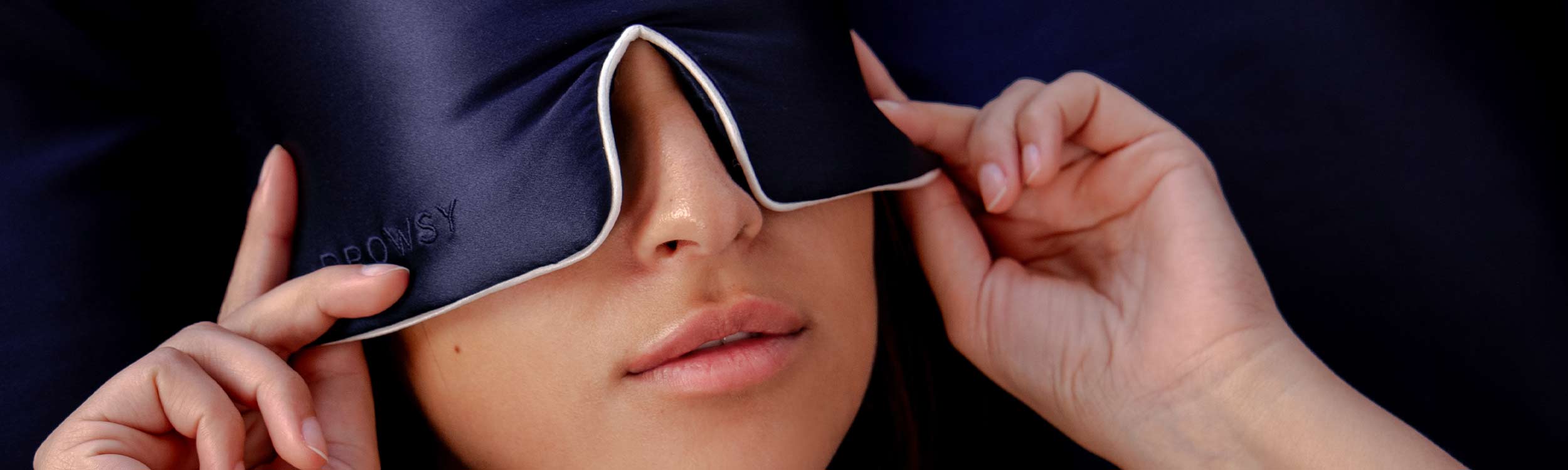 A person adjusts a dark blue sleep mask over their eyes, with their hands on either side of the mask, partially covering their face and lips.