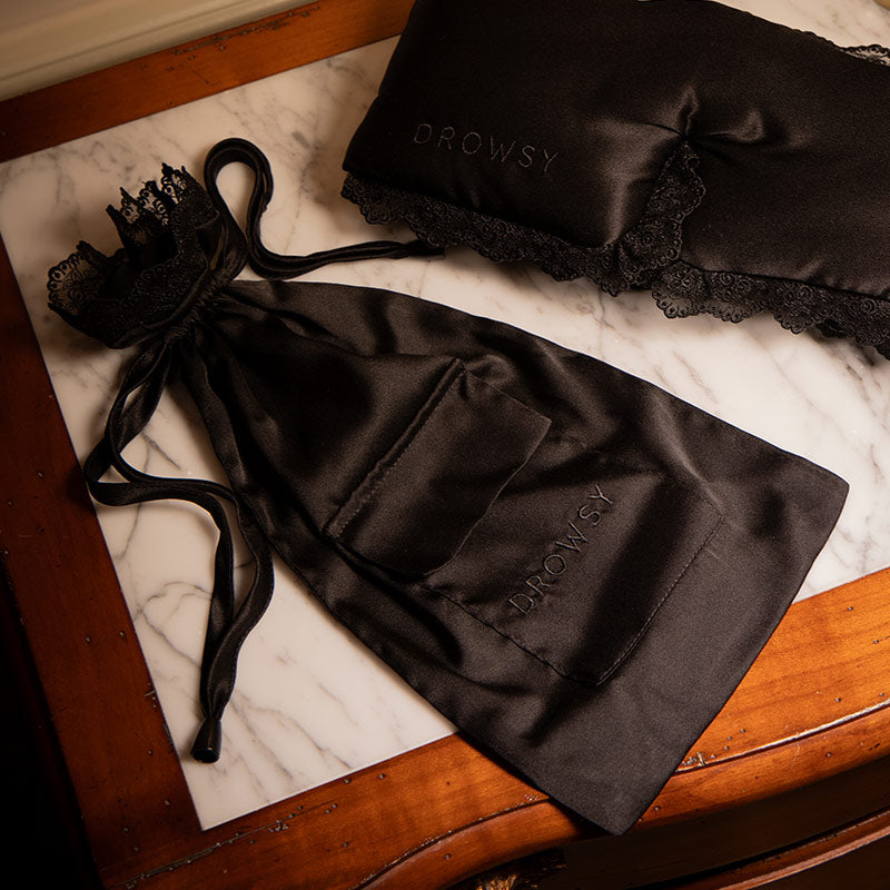A black silk sleep mask with lace trim and "DROWSY" embroidery by drowsysleepco rests beside the matching Paris Love Story Silk Carry Pouch on a marble-and-wood surface.