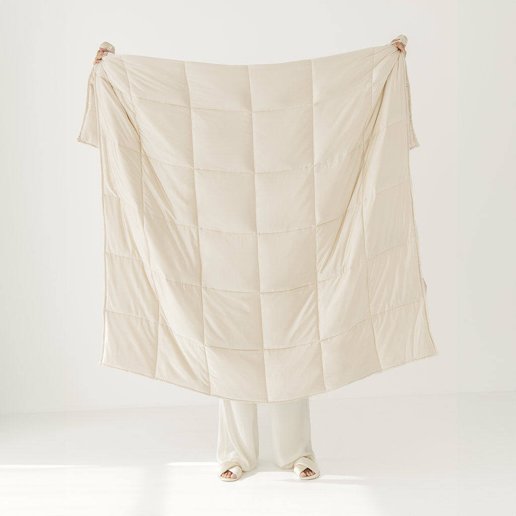A person holds up the Dusty Gold Silk Weighted Blanket by us.drowsysleepco, a luxurious, cream-colored blanket designed to cover your whole body and provide deep relaxation for an unrivaled sleep, set against a bright, minimalistic white room.