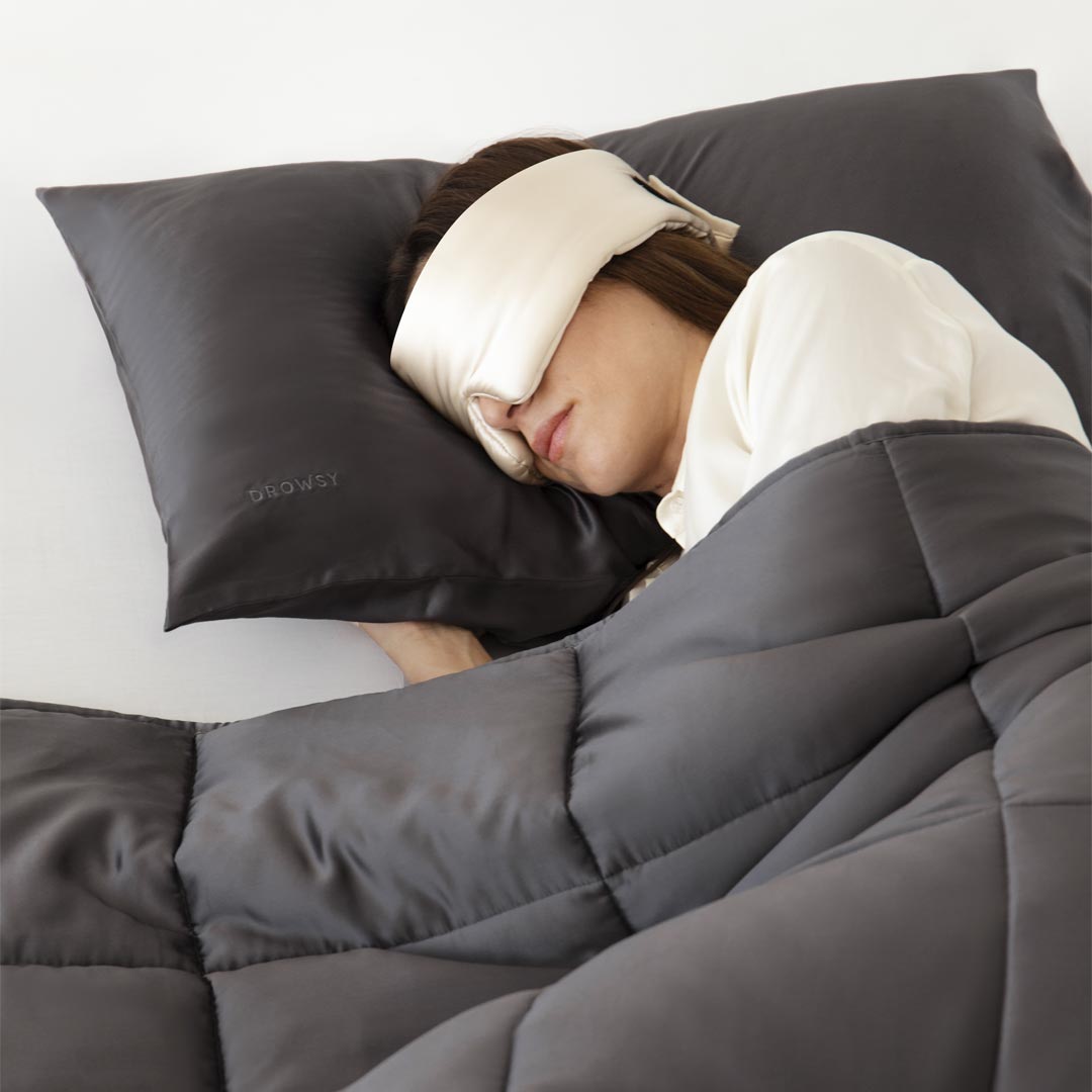 A person sleeps on a gray pillow under a gray quilted blanket, wearing a white sleep mask.