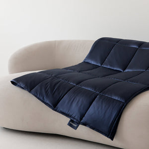 The Midnight Blue Silk Weighted Blanket by us.drowsysleepco is draped over a modern curved light beige sofa against a white wall, with a small fabric tag visible on the blankets edge.