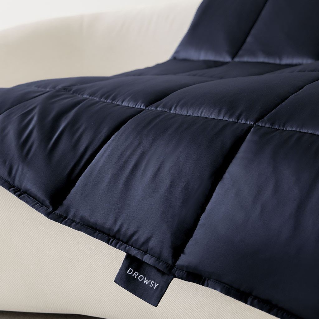 The us.drowsysleepco Midnight Blue Silk Weighted Blanket, featuring a smooth, soft silk cover and quilted design, is draped over a light beige surface. The dark navy color and Drowsy label add a touch of elegance.