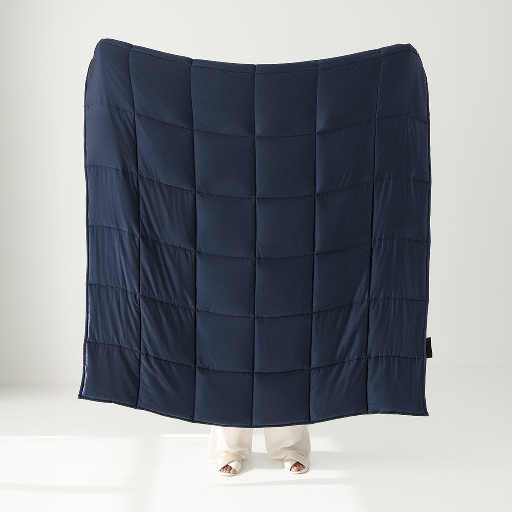 A person holds up the us.drowsysleepco Midnight Blue Silk Weighted Blanket, with only their lower legs and feet visible beneath it. The plain white room background makes the luxurious navy blue blanket stand out.