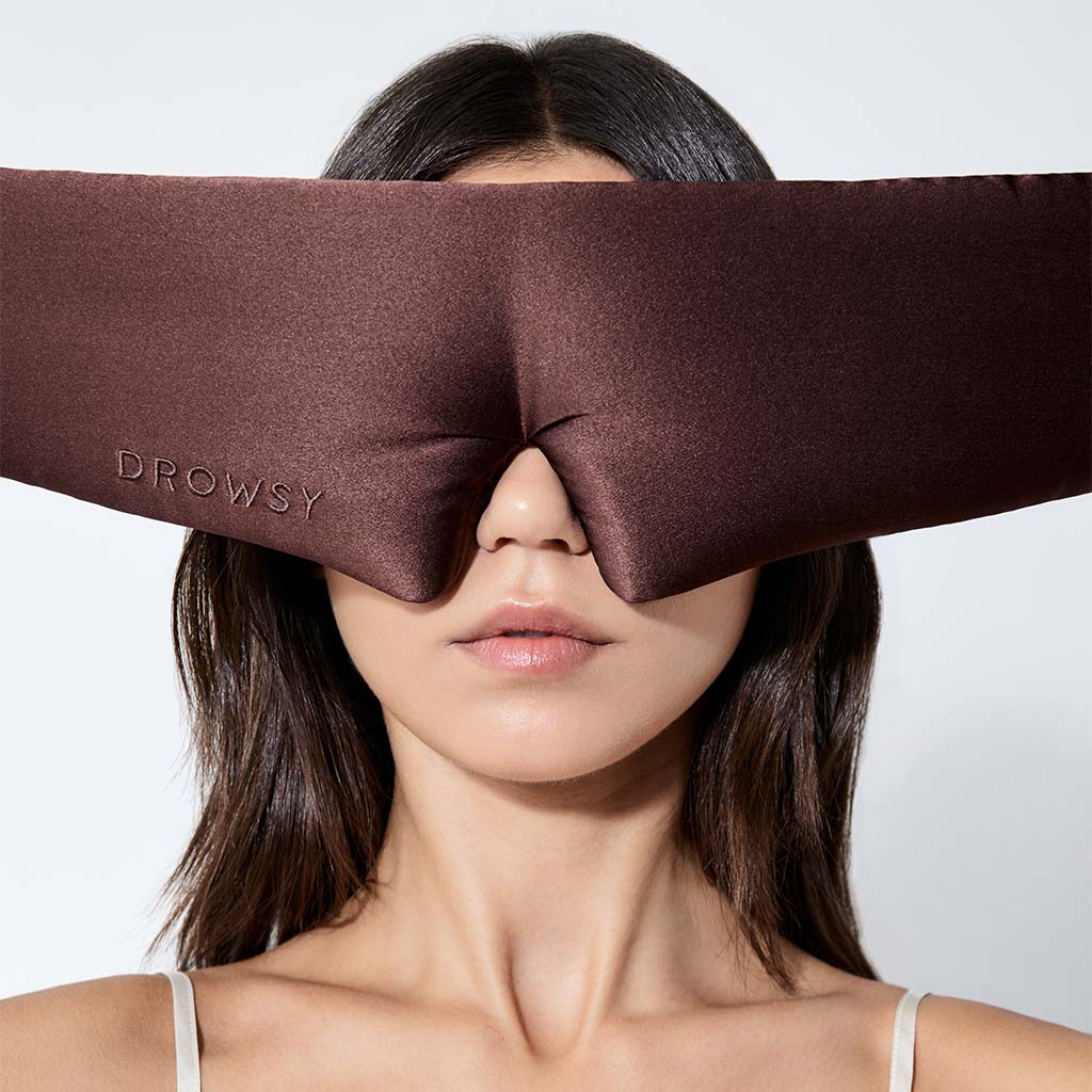 A woman with straight brown hair wears a large brown silk sleep mask labeled "Cocoa Glow" by drowsysleepco, covering her eyes and upper nose. She faces forward against a plain white background, showing comfort and style.