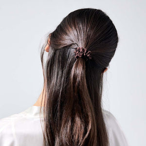 A woman with long, straight brown hair, seen from behind in a white top, styles her hair using the drowsysleepco Skinny Scrunchie Set - Cocoa Glow for gentle and protective hold.