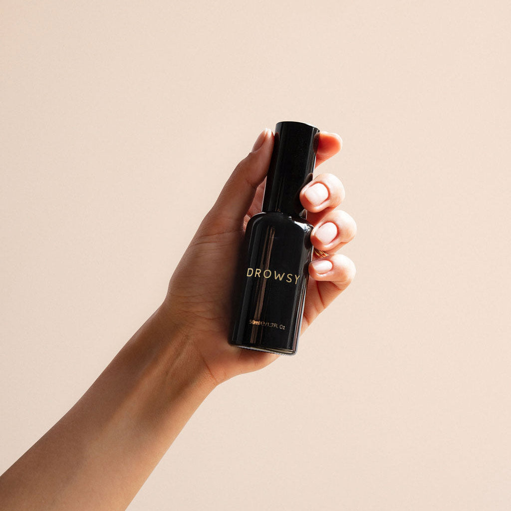 A hand holds a black Sleep S.O.S Pillow Spray bottle by us.drowsysleepco, infused with natural oils, against a light beige background.