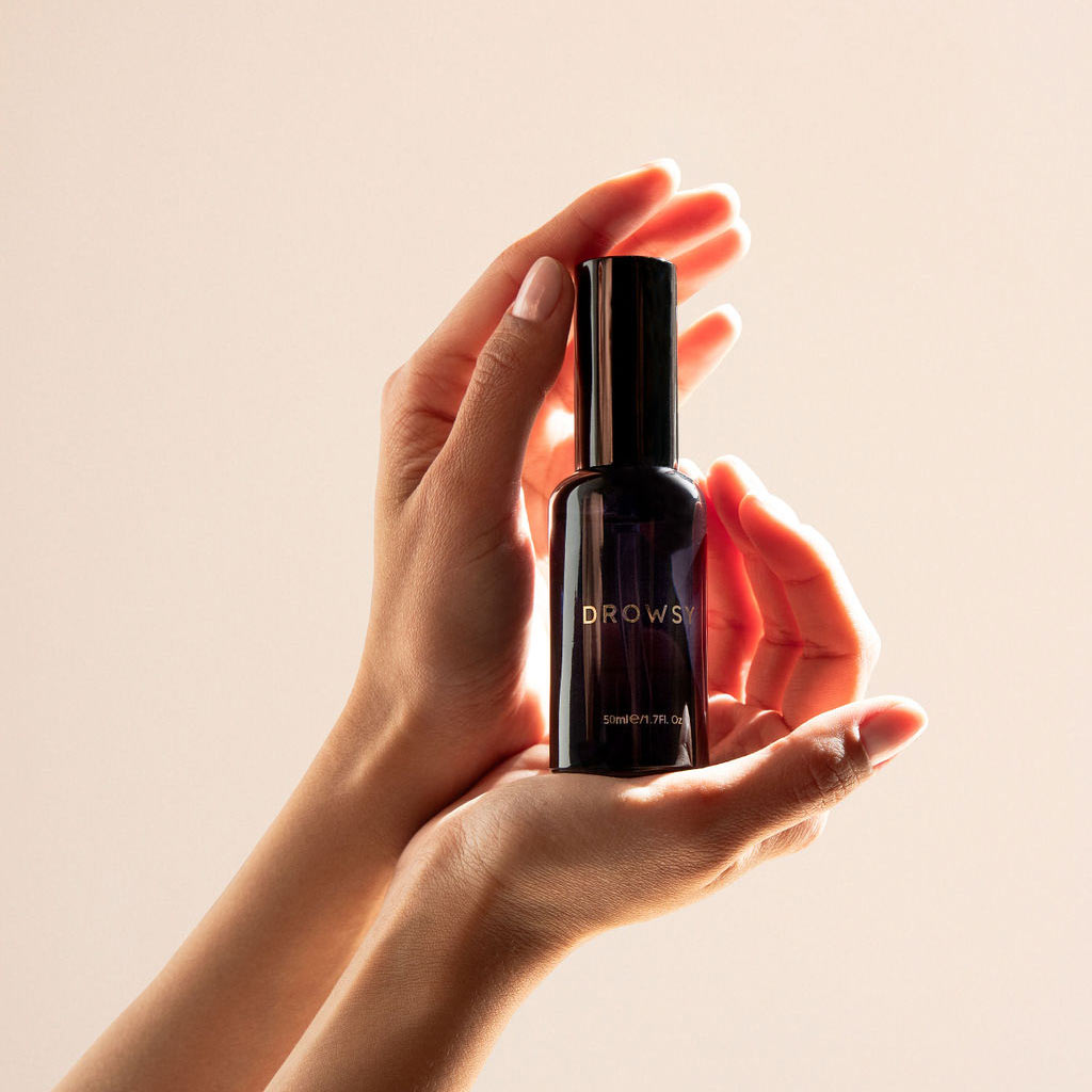 A pair of hands gently holds a dark-colored bottle labeled Sleep S.O.S Pillow Spray by us.drowsysleepco, infused with natural oils, against a softly lit background—inviting restful sleep.