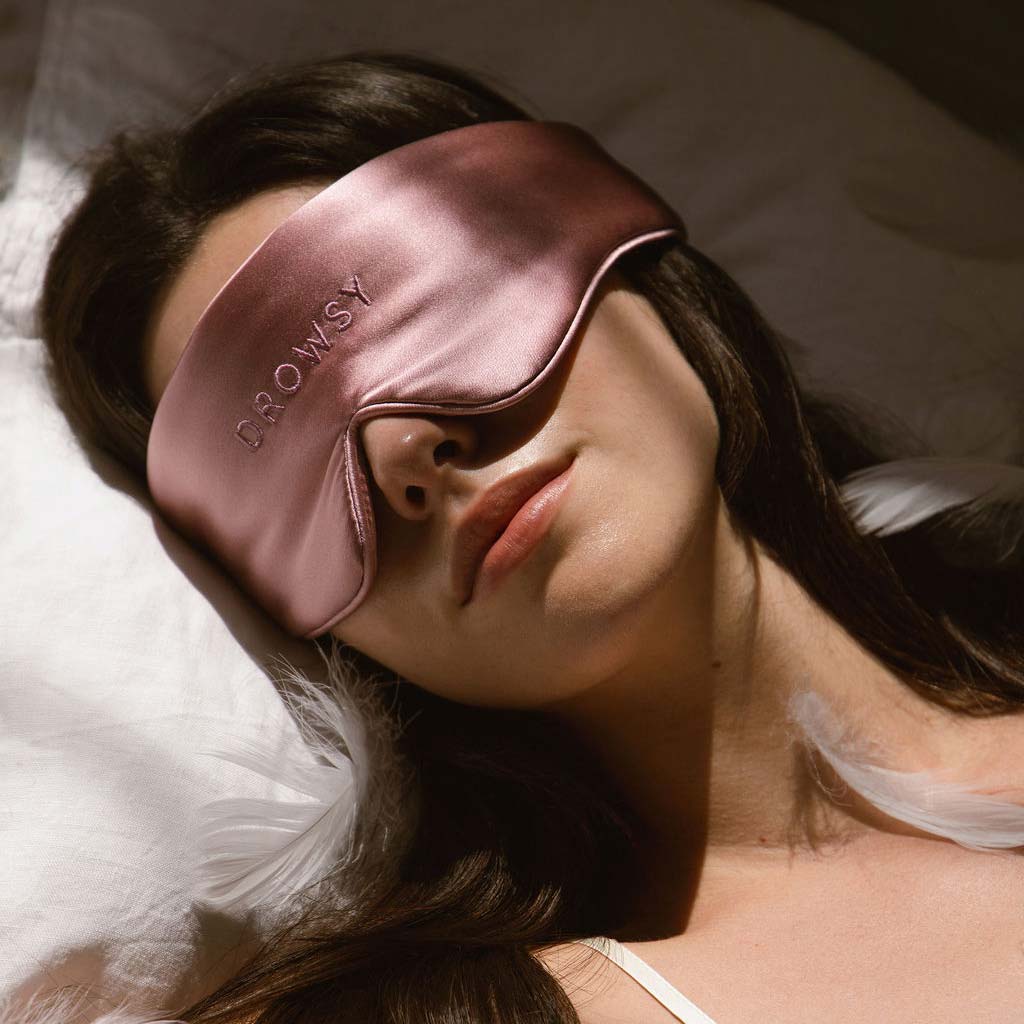 A woman lies on her back in bed wearing a silky pink sleep mask that says DROWSY. She has long dark hair and is dressed in a white top, resting on a white pillow with soft lighting.
