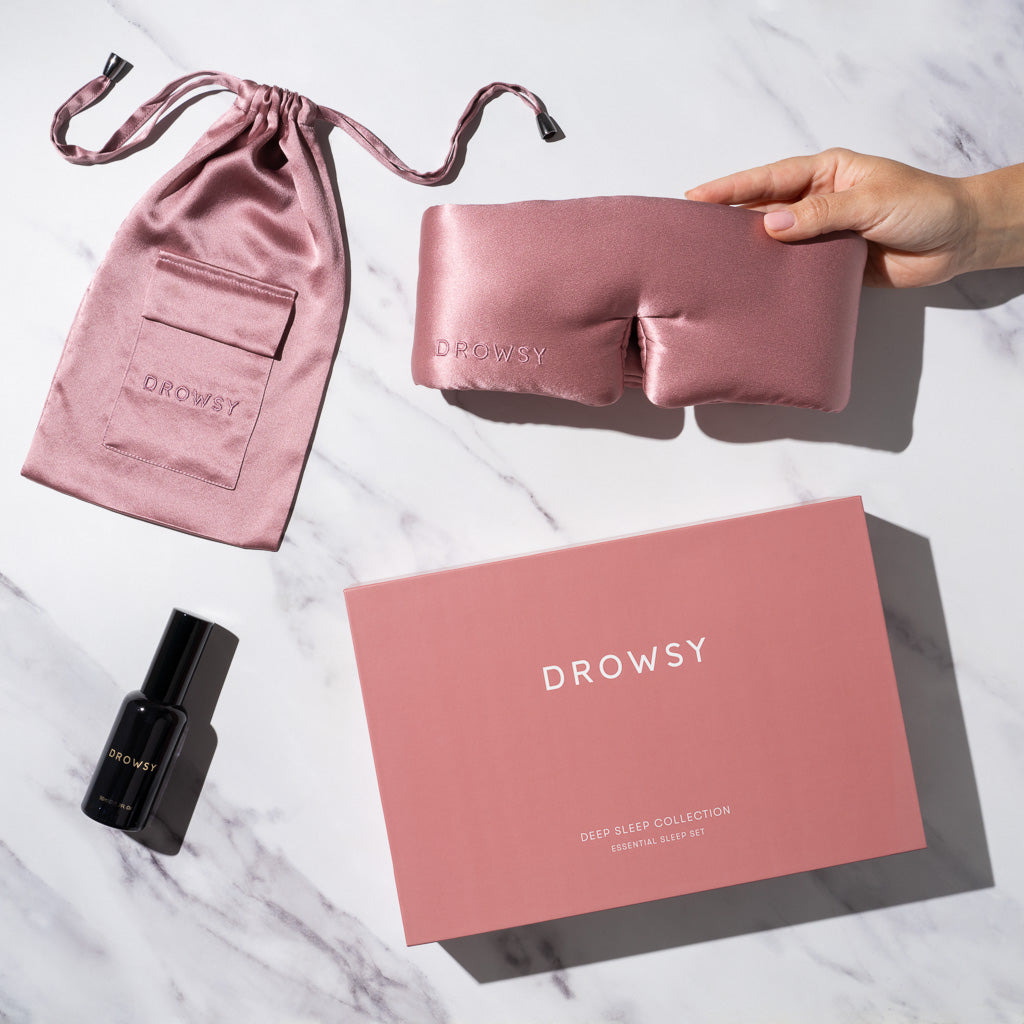 The Damask Rose - Deep Sleep Collection by us.drowsysleepco features a luxury silk sleep mask, matching pouch, black deep sleep pillow spray, and pink box—essentials for your nightly routine, all arranged on a white marble surface.