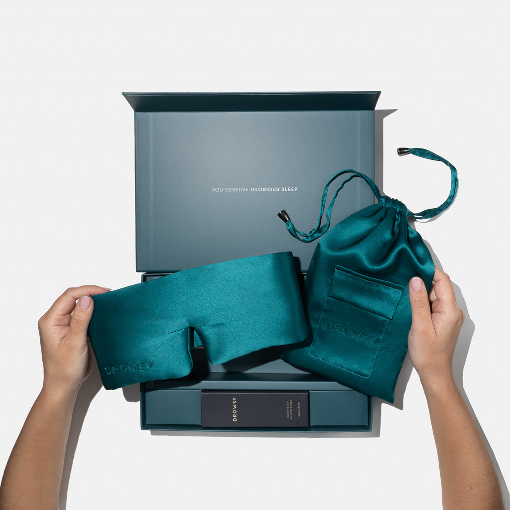 A person holds a Green Sapphire - Deep Sleep Collection silk sleep mask and drawstring pouch by us.drowsysleepco above an open gift box filled with sleep essentials, with "YOU DESERVE GLORIOUS SLEEP" printed inside the lid.