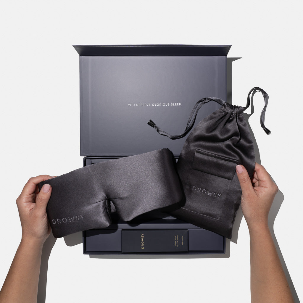 Two hands hold the Moonlight Shadow - Deep Sleep Collection by us.drowsysleepco: a luxury silk sleep mask and pouch above a gift box with deep sleep pillow spray. Inside the lid reads "YOU DESERVE GLORIOUS SLEEP.