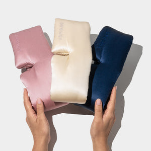 Two hands hold three Drowsy Besties Pack luxury masks in pink, cream, and navy blue. Crafted from mulberry silk and embossed with the brand name, they sit side by side on a light background—perfect for beauty sleep.