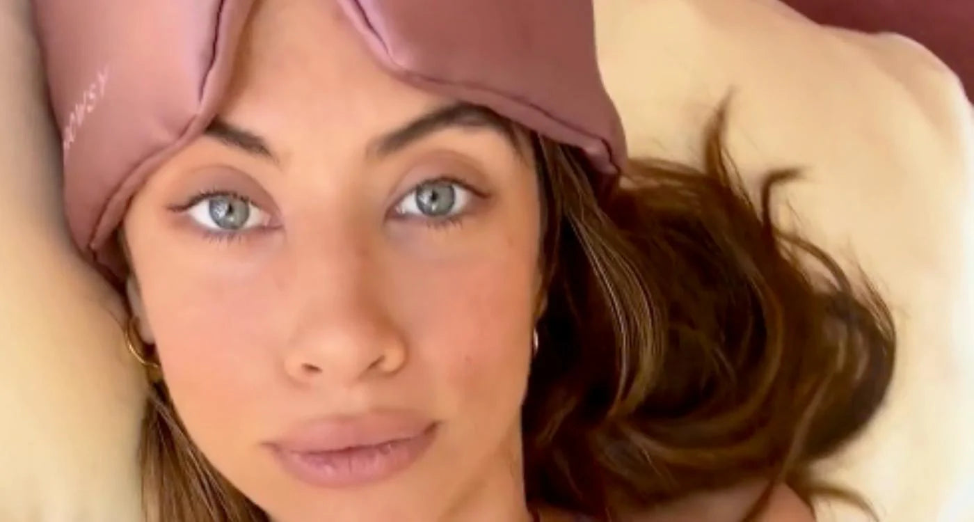 A woman with light eyes lies on a pillow, wearing a pink sleep mask pushed up on her forehead. She has long brown hair and looks directly at the camera with a relaxed expression.
