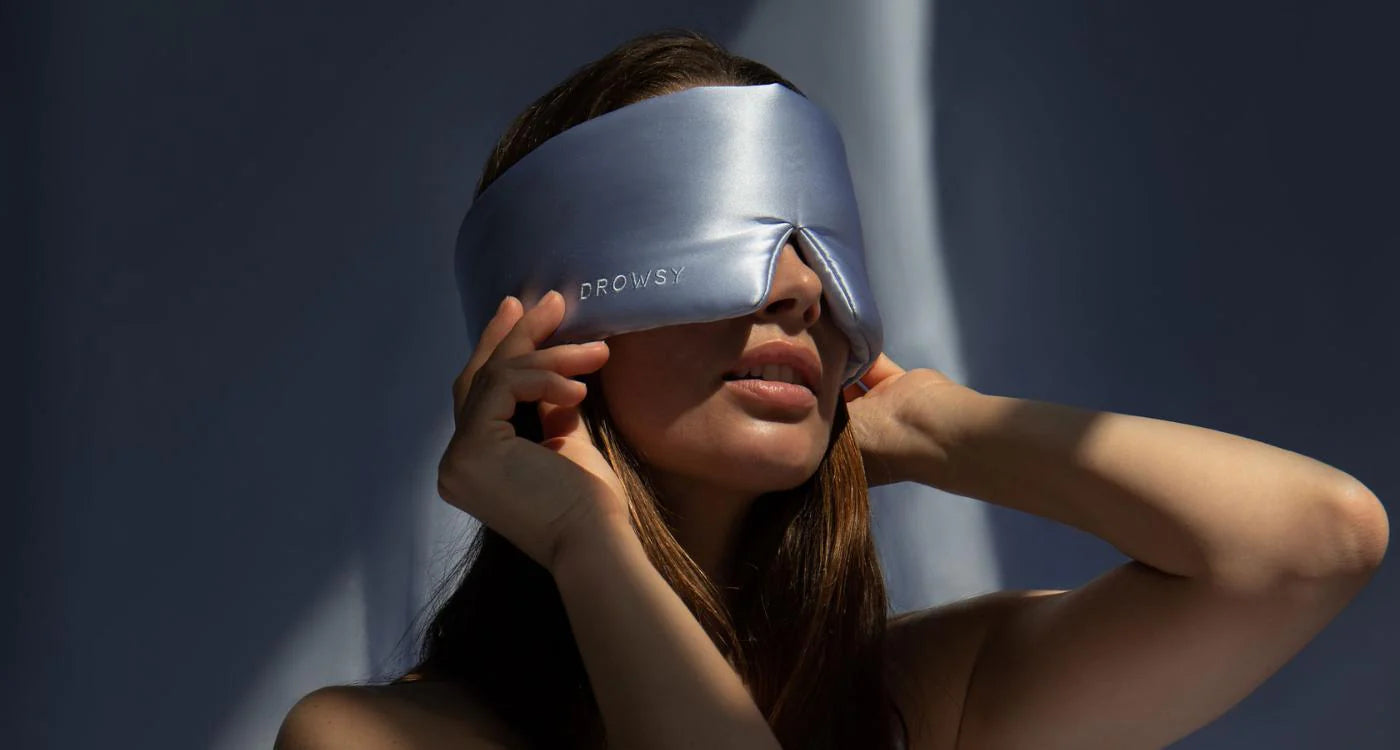 A woman adjusts a large, light blue silk sleep mask labeled DROWSY over her eyes, with soft shadows and gentle lighting in the background.