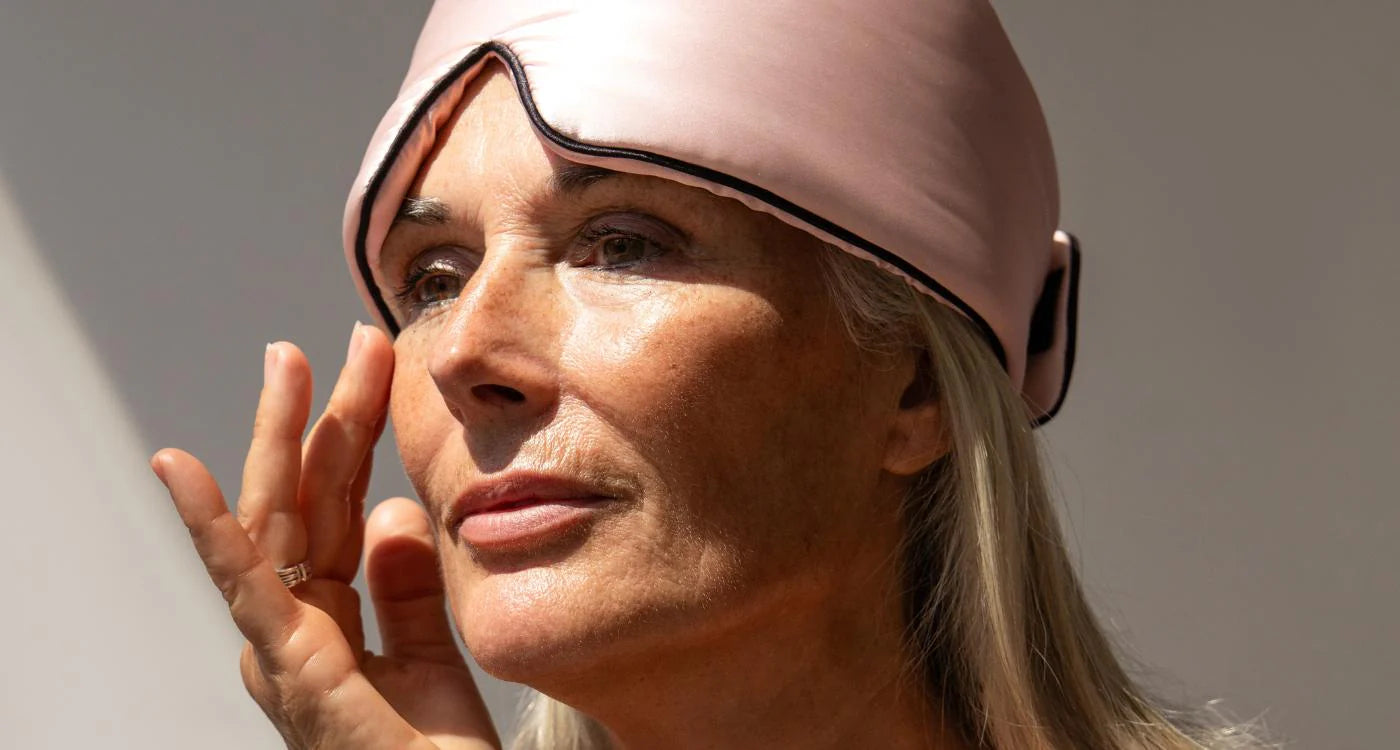 A woman with long gray hair wears a pink satin sleep mask pushed up on her forehead and gently touches her face while looking thoughtfully into the distance.