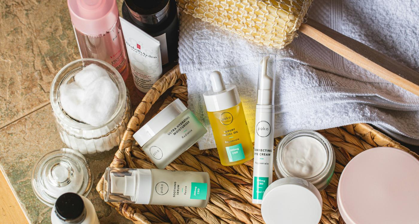 A variety of skincare products, including creams, serums, and oils, are arranged in and around a wicker tray with cotton pads, a towel, and a bath brush on a tile surface.