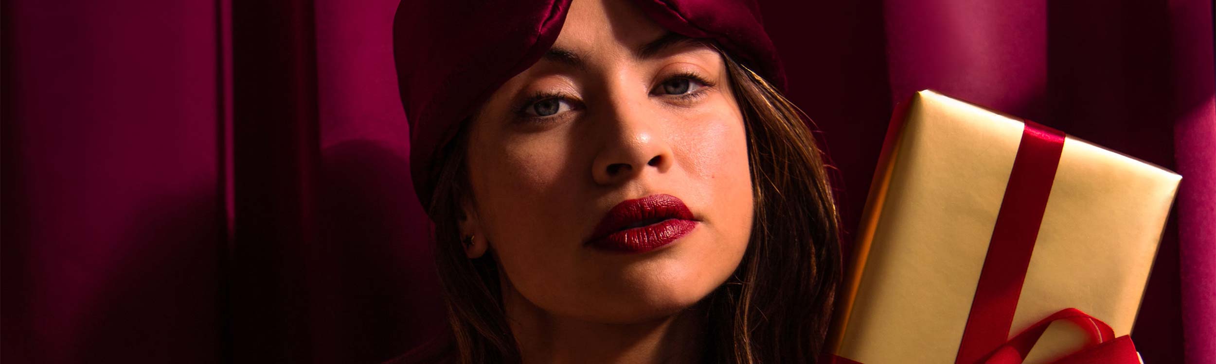 A woman wearing a burgundy eye mask and lipstick holds a gold gift box wrapped with a red ribbon, standing in front of matching burgundy curtains.