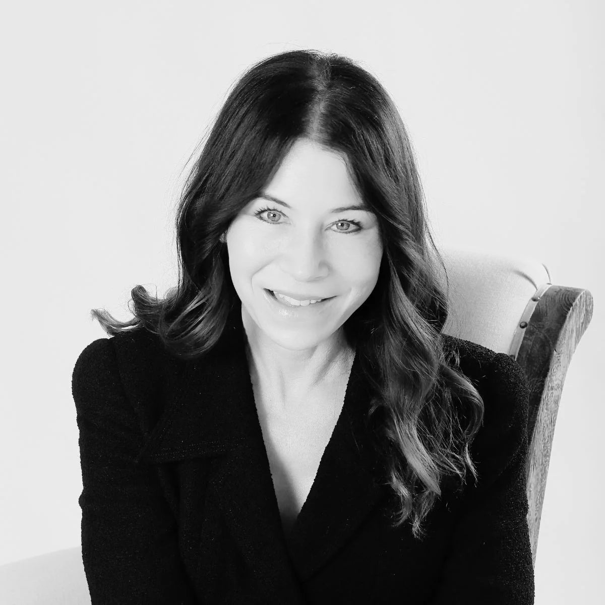 Black and white portrait of a woman with long, wavy dark hair, wearing a dark jacket and smiling while seated on a light-colored chair against a plain background.