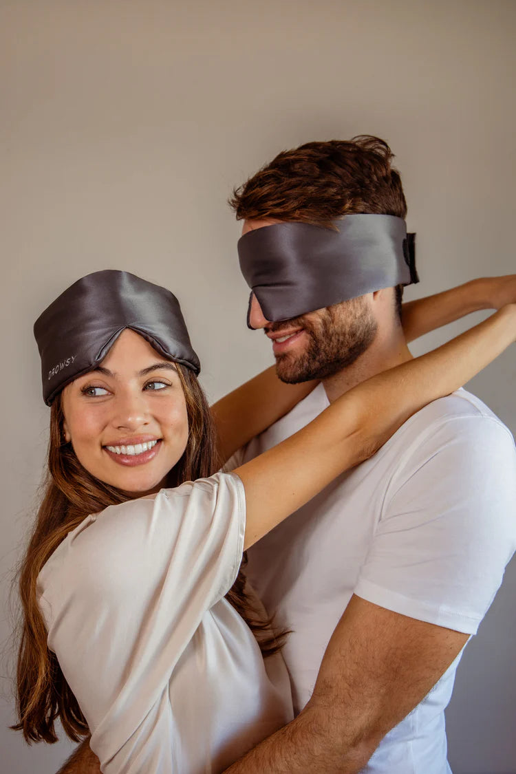 A smiling woman with long brown hair hugs a man from behind. Both wear dark gray sleep masks on their heads; hers is above her eyes, his covers his eyes. They are dressed casually in light-colored clothing.