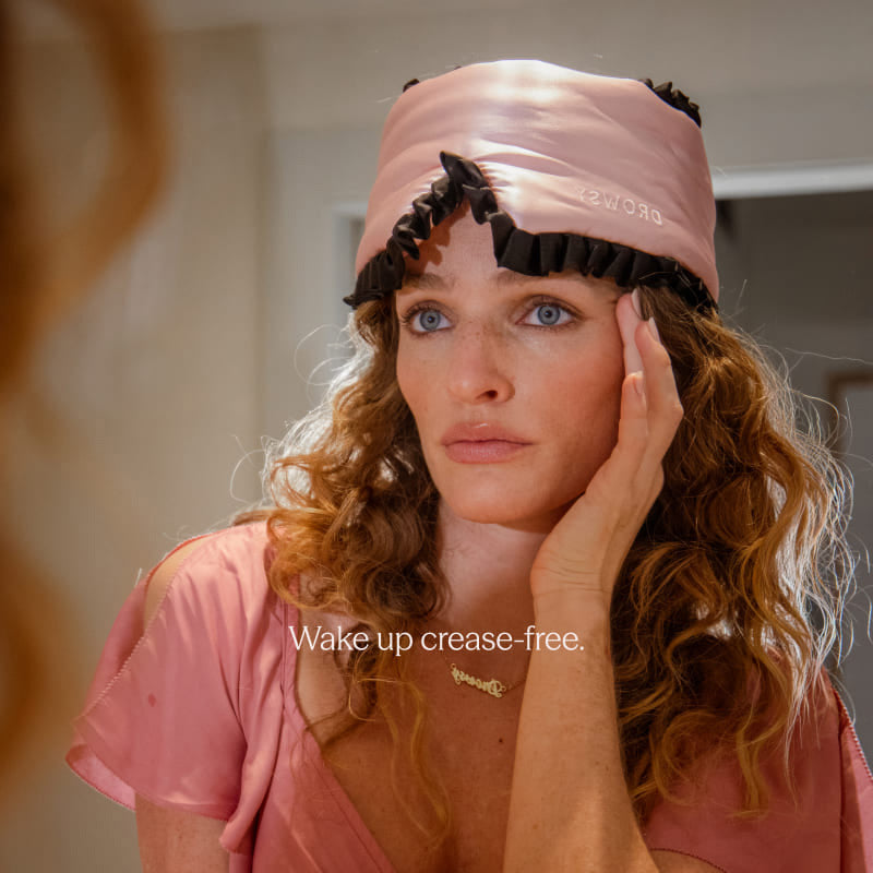 A woman with wavy hair wears a drowsysleepco Sleep and the City Pink & Black mulberry silk sleep bonnet and matching pajamas as she looks in the mirror. Text on the image reads: Wake up crease-free.