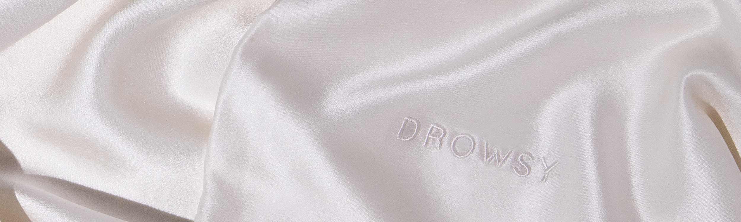 Close-up of smooth, shiny, light-colored satin fabric with the word DROWSY embroidered in matching thread on the surface.