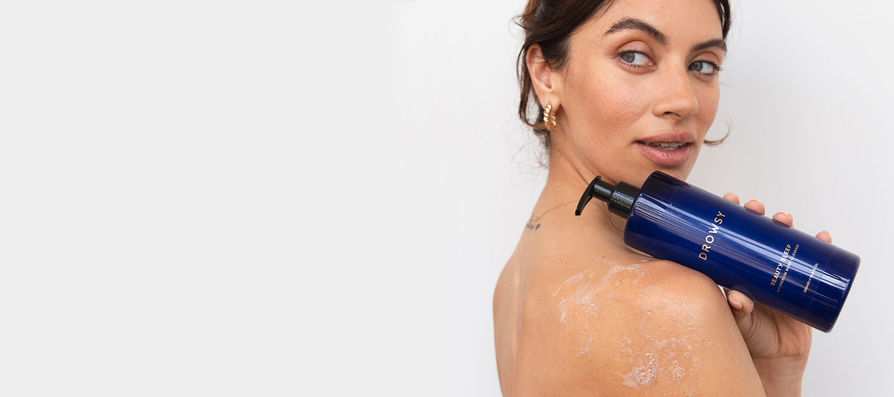 A woman with glowing skin looks over her shoulder while holding a blue bottle of Drowsy Sleep Zen Body Wash; some of the product is visible on her shoulder.