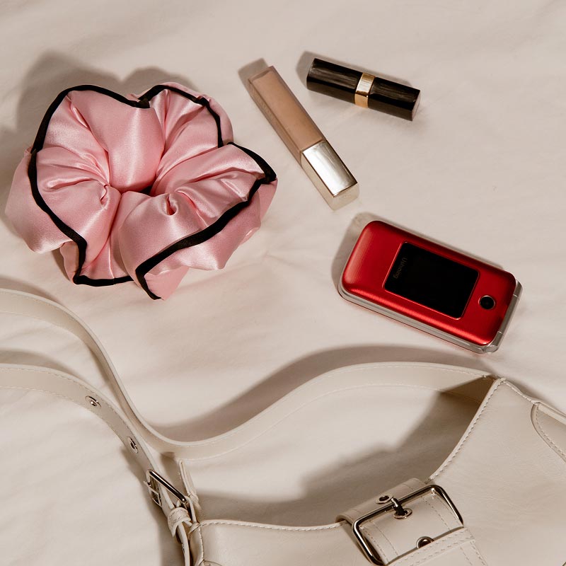 A drowsysleepco Sleep and the City Pink Padded Scrunchie, concealer, lipstick, red flip phone, and part of a white handbag with a buckle are arranged on a beige surface.