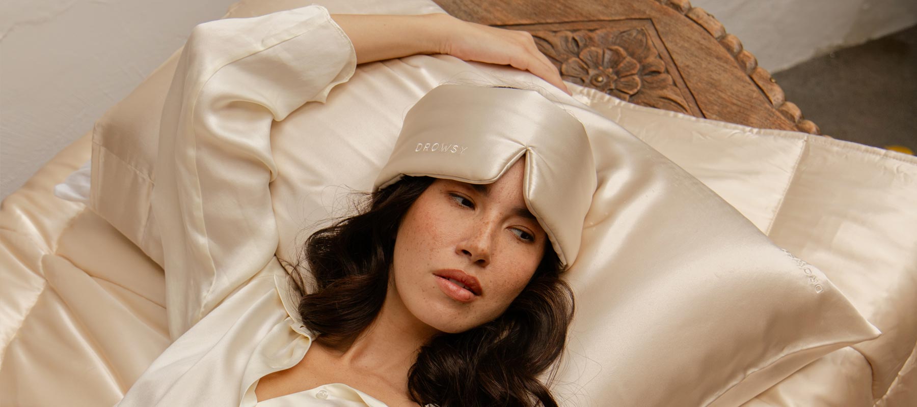A woman with long dark hair rests on a bed with cream-colored satin sheets, wearing a matching satin sleep mask and robe, with one arm above her head on a carved wooden headboard.