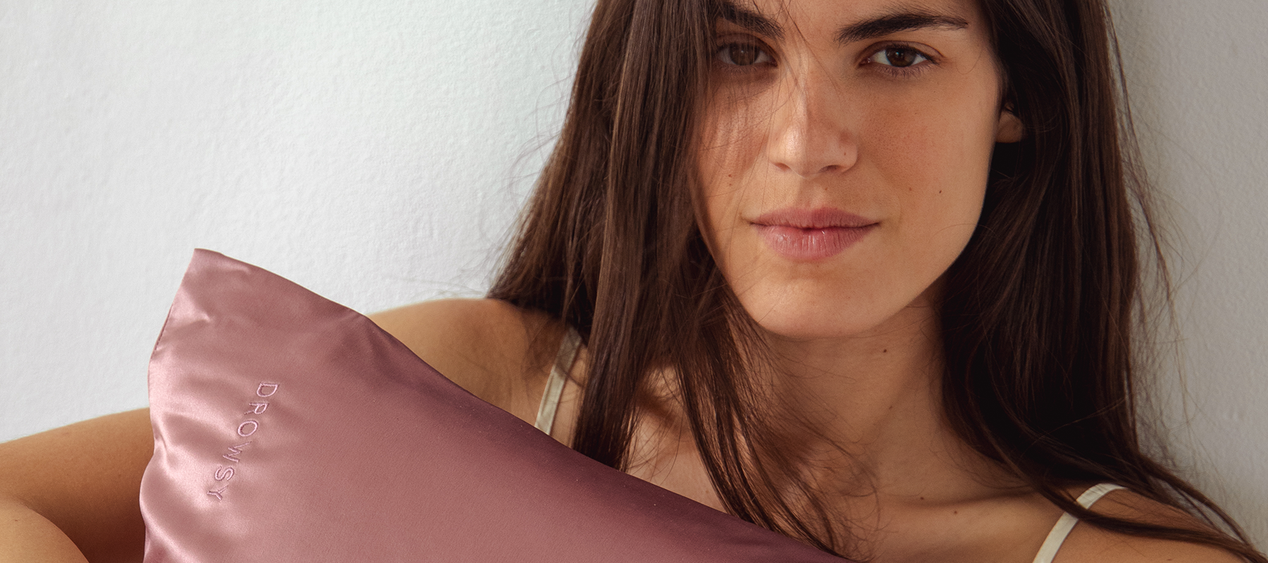 A woman with long brown hair holds a mauve satin pillow embroidered with the word DROWSY, looking softly at the camera against a plain white background.