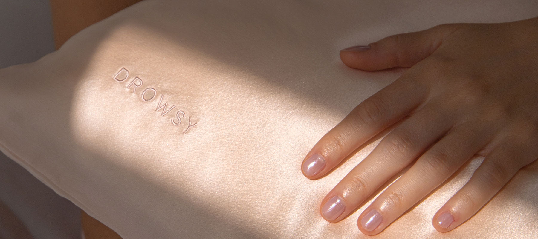 A hand with neatly manicured nails rests on a light pink silk pillow embroidered with the word DROWSY. Sunlight creates soft shadows across the fabric and hand.