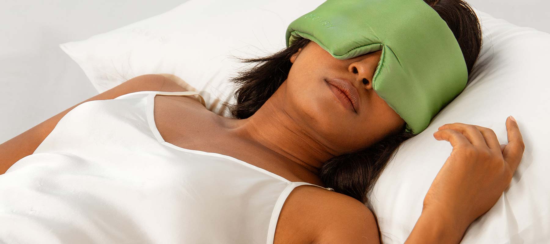 A woman in a white camisole lies on a white pillow with a large green sleep mask covering her eyes, appearing relaxed and restful.